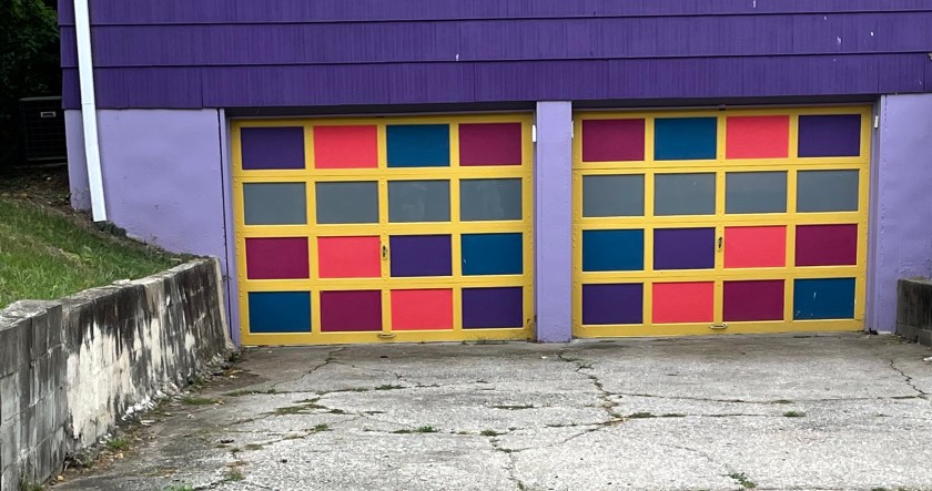 Two side-by-side garage doors painted in squares of several vibrant colors. The surrounding house walls are shades of purple.