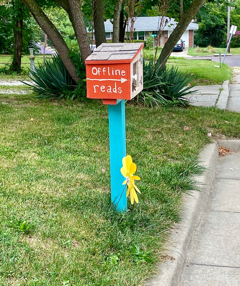A little free library, wooden red container with the words "offline reads" painted in white, mounted on teal post. A plastic yellow spinning flower is in the ground next to it.