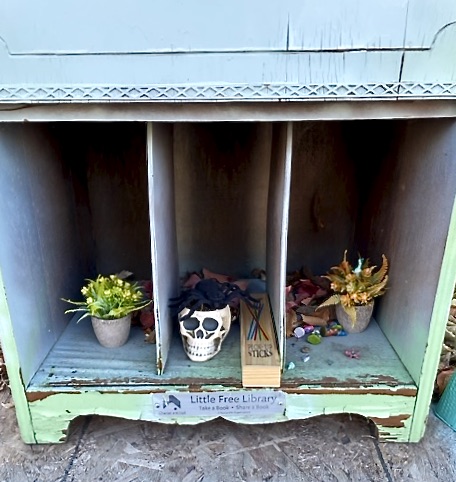 Old wooden cabinet, blue and green, with two plants, a fake skull topped by a fake spider, some pick-up sticks, and miniature people.