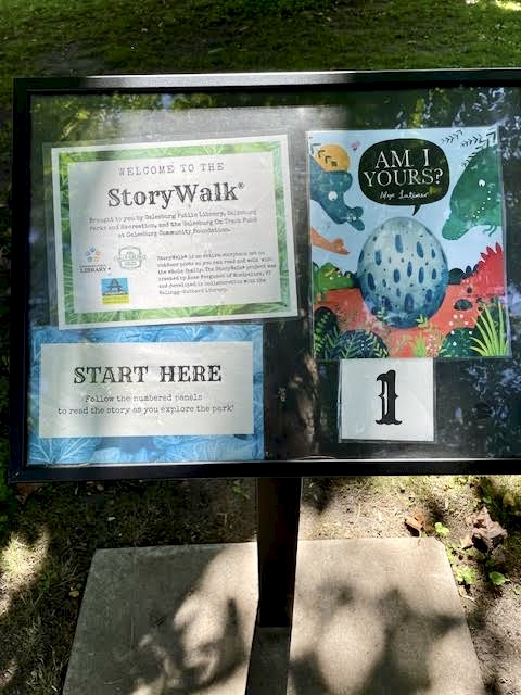 Metal stand with signs under glass: "Story Walk...Start Here" and a book cover "Am I Yours?" Alex Latimer