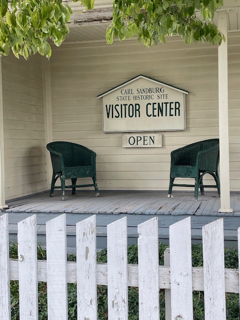 Two wicker chairs on a blue porch on either side of a sign saying Carl Sandburg State Historic Site Visitor Center...open