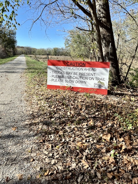Sign on trailside reads "Caution, construction ahead, trucks may be present, please use caution on trail, please slow down"