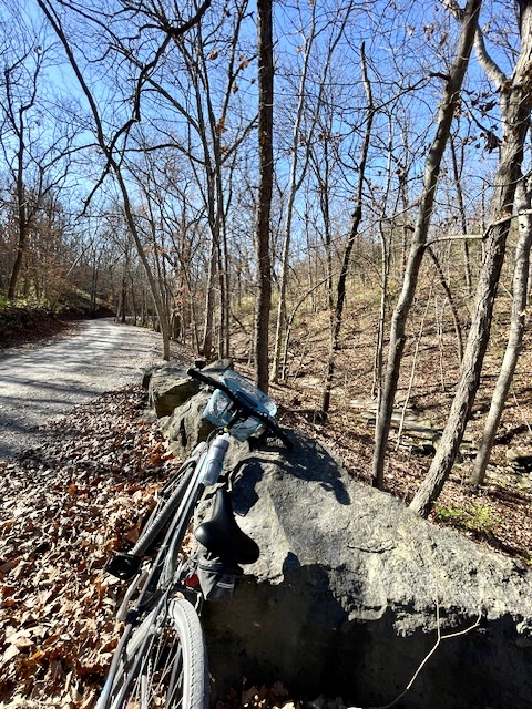 Bicycle in foreground leaning against rocks. Gravel trail running into the distance with trees on either side.