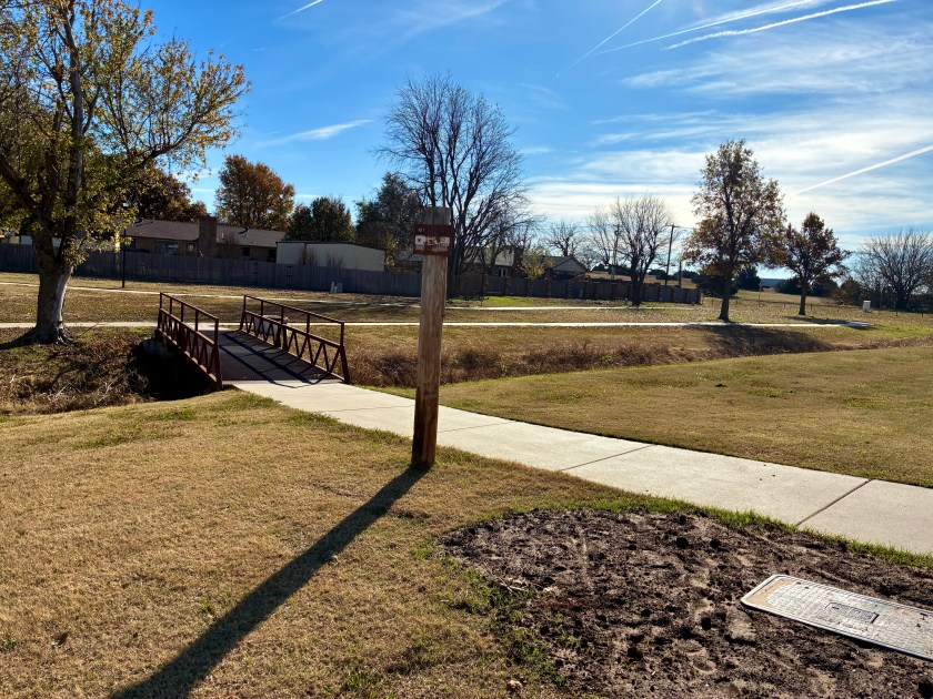 Footbridge over ditch leading to cement path in park. Building in background.