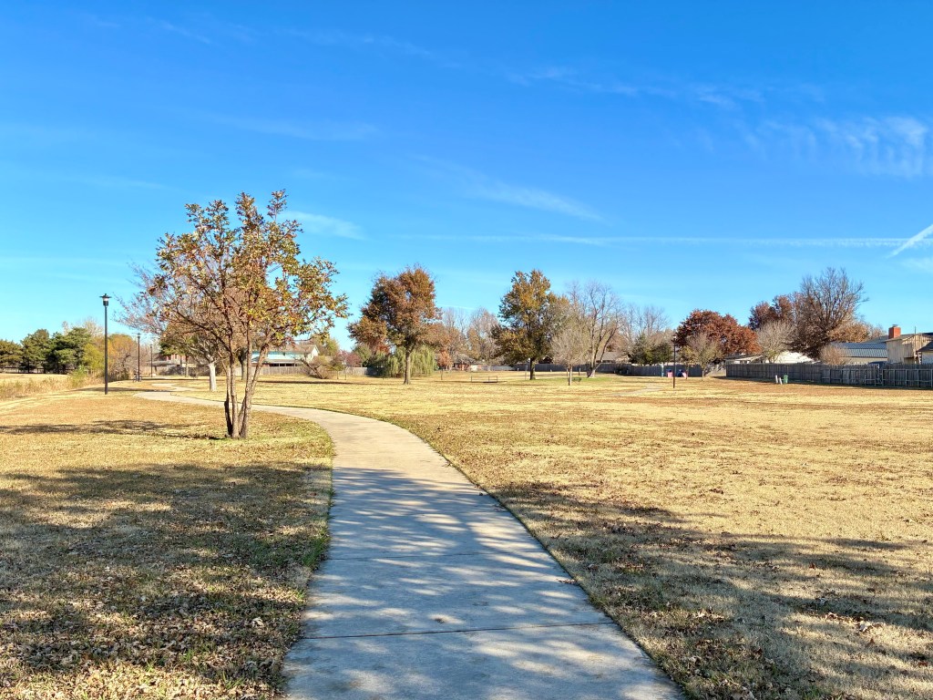 Cement walkway through park. Small trees here and there. Blue sky.